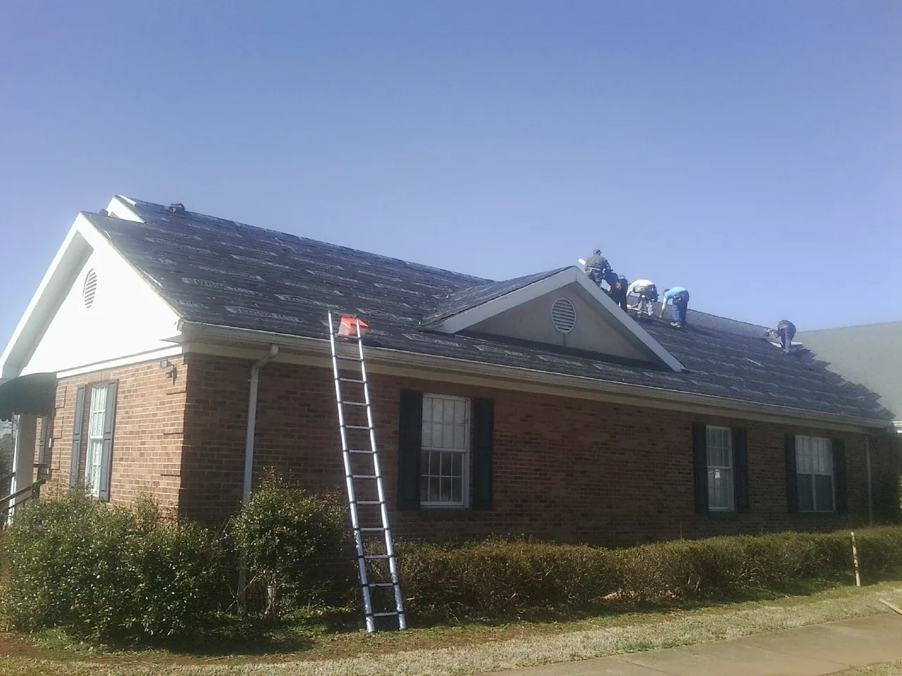 Crew tearing shingles off a brick ranch roof with a ladder set — Emergency Roof Repair work by our crew in Lula