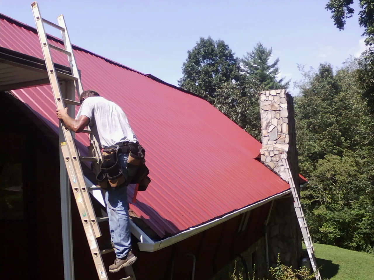 Roofer on a ladder beside a red metal roof and stone chimney — Soffit & Fascia Repair by Roofing Brothers in Lula